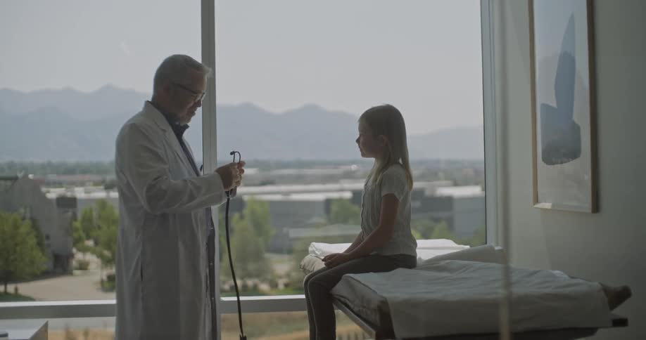 Silhouetted against a large window with a mountain view a friendly doctor talks to a young girl as he prepares his stethoscope for an examination