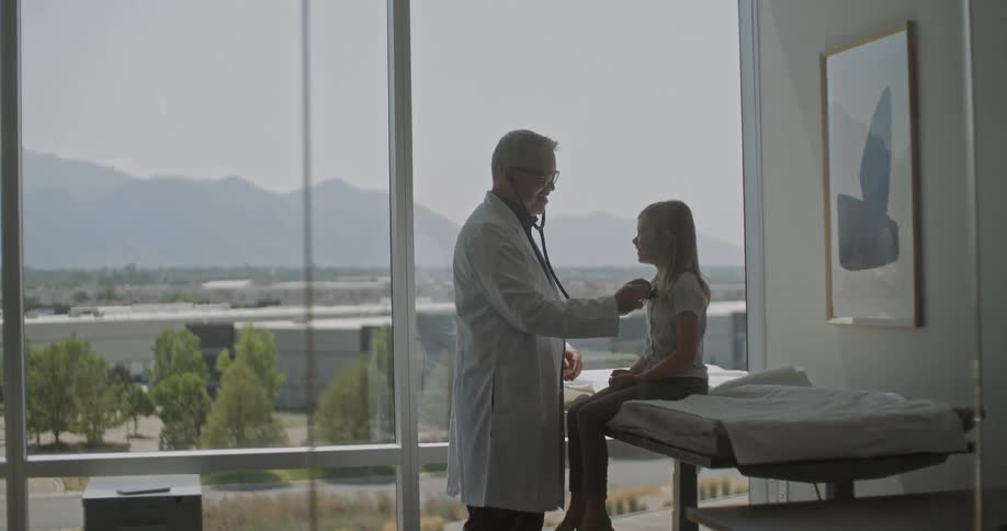 A doctor silhouetted against a large window with a mountain view uses a stethoscope to examine a young girl sitting on an examination table