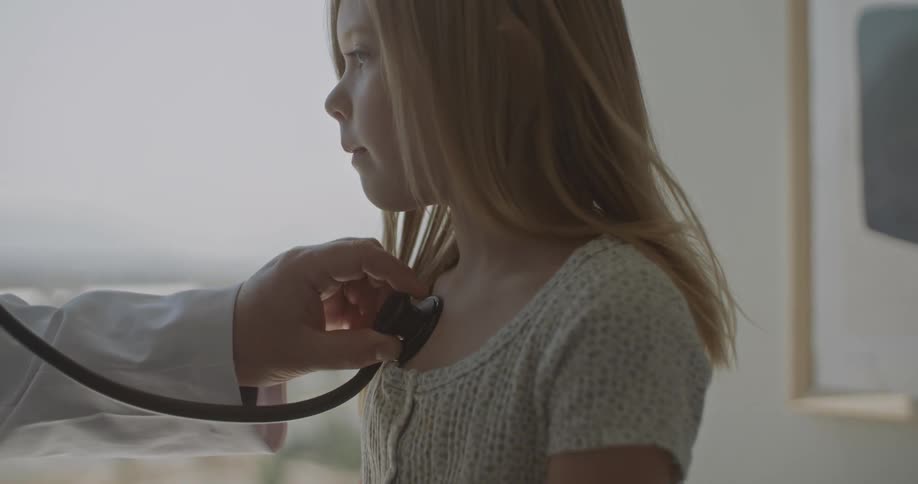 A doctor places a stethoscope on a young girls chest during a medical examination in an office with a large window