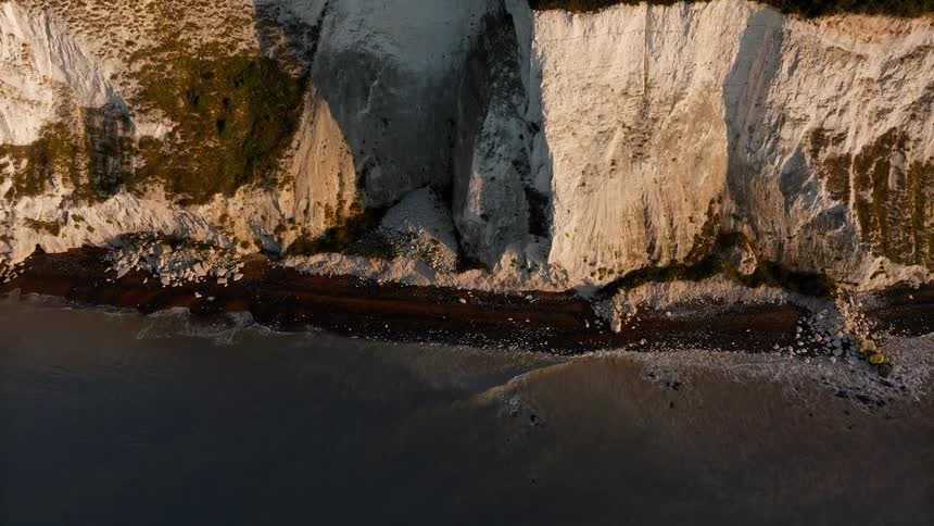 An aerial drone shot moves toward a massive section of white chalk cliffs where a recent rockfall has deposited a pile of debris onto the dark beach below while small waves lap at the shoreline