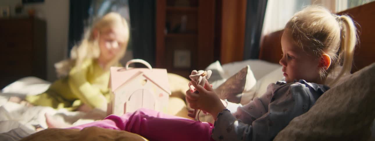 Two young girls play quietly on a sunlit bed with one child carefully examining a small doll while the other interacts with a toy house in the background
