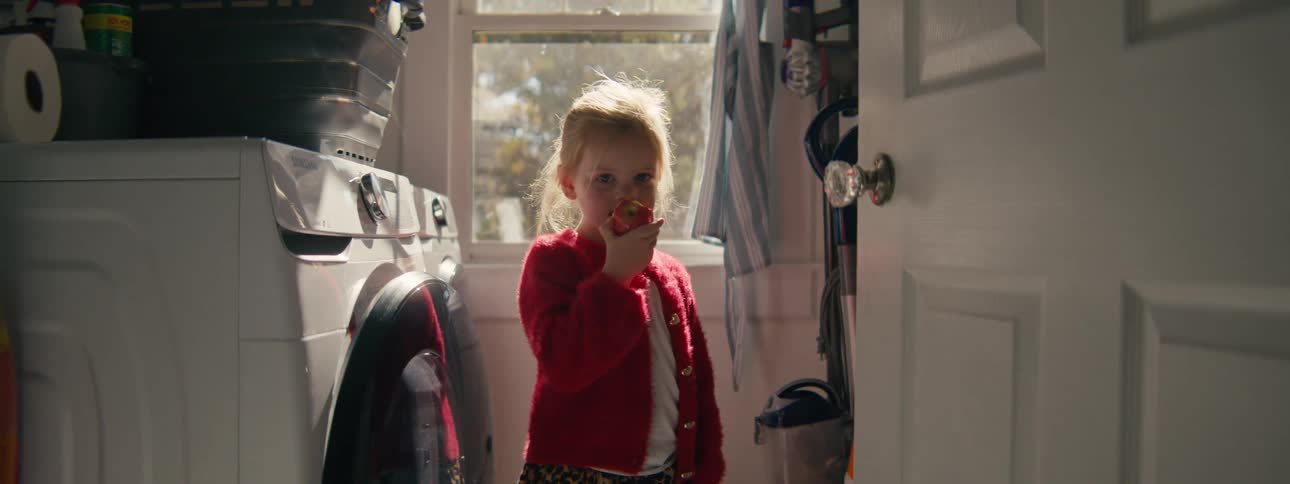 A young child eats an apple while standing in a softly lit laundry room beside a washing machine