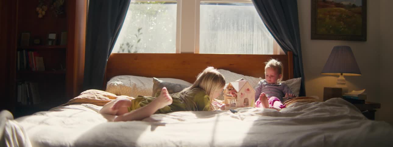 A medium view shows two young girls playing with a small wooden dollhouse and figurines while sitting on a large bed in a sunlit room