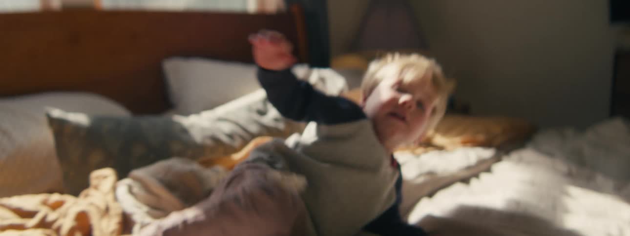 A young boy jumps energetically on a sundrenched bed before collapsing onto the messy blankets with a joyful expression as morning light streams through the bedroom window