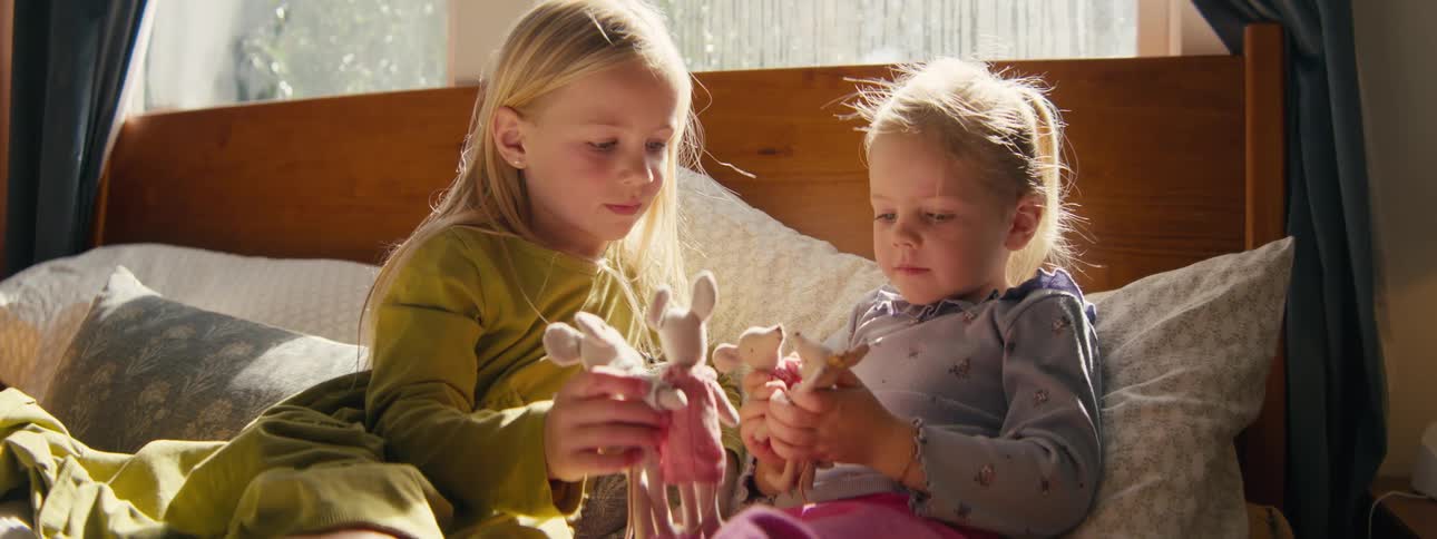 Two young girls play with small dolls while sitting together on a bed in warm daylight