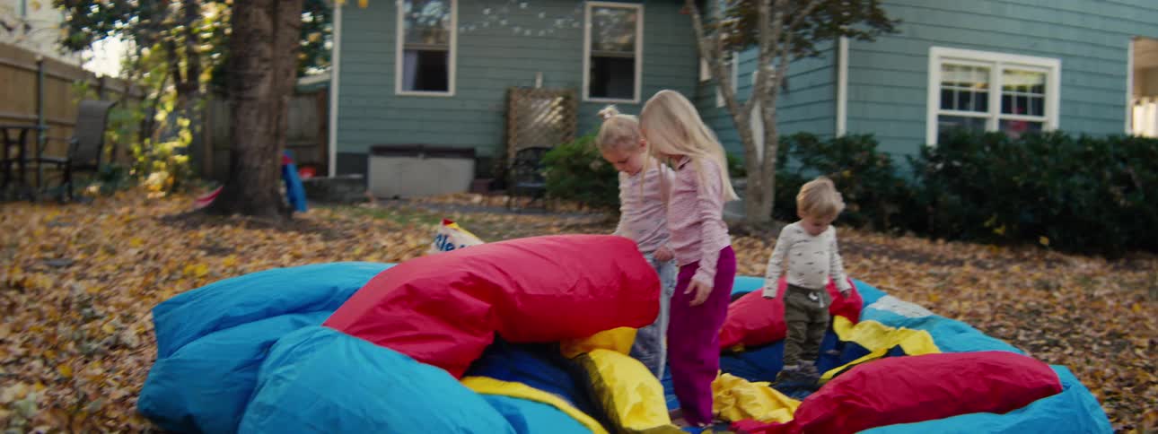 Children jump on a colorful inflatable toy in a residential backyard covered with dry leaves