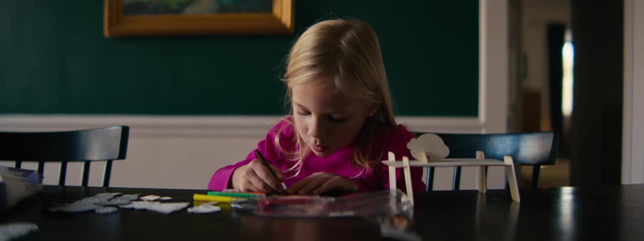 A young girl focuses on drawing and crafting at a dining table inside a home during daylight