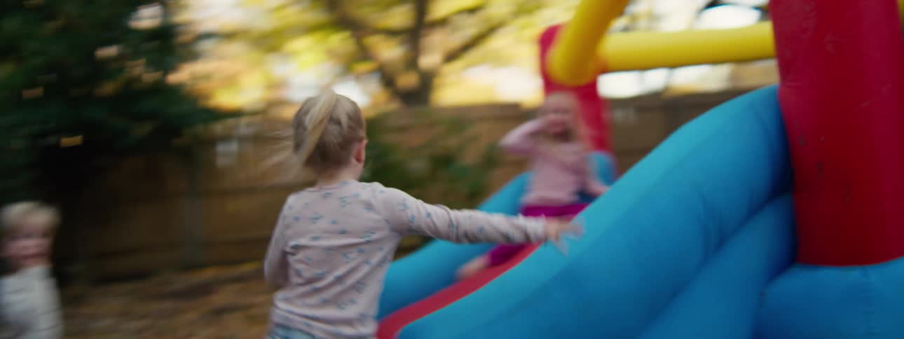 A young girl runs through a backyard covered in autumn leaves and climbs onto a colorful inflatable bounce house to play with another child and the camera follows her from behind as she excitedly joins other girl on the red and blue jumping surface