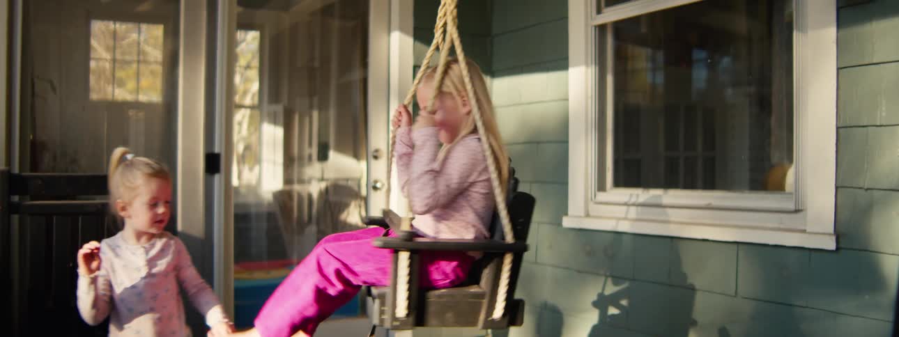 Little girl sitting in a swing on the front porch spinning around as her little sister watches