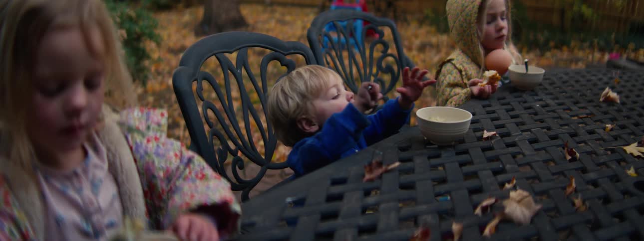 Children seated at an outdoor table eating from bowls with fallen leaves scattered and chairs around