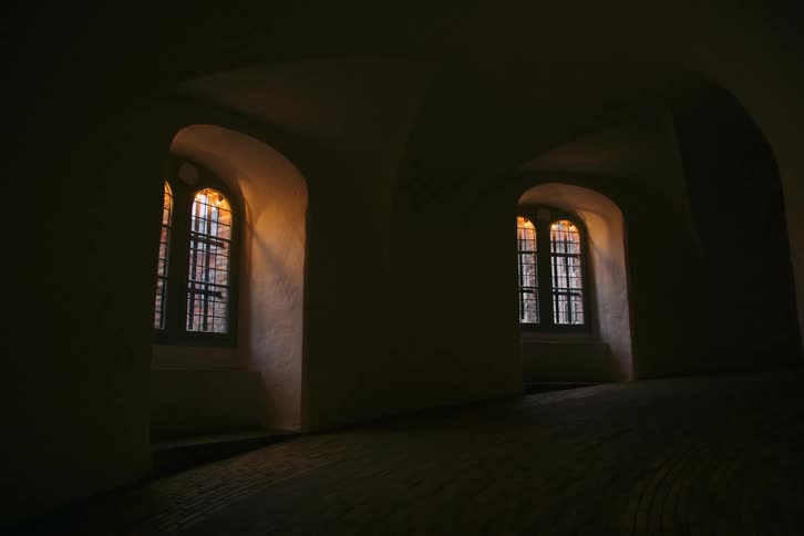 Interior corridor with arches and tall windows illuminated by warm light in a historic building