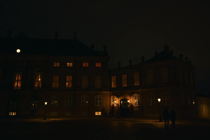 This video shows a large historic building at night with warm light glowing from its many windows two people are seen walking across the dark courtyard in front of the illuminated entrance