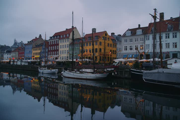 Boats docked in the canal along the coast as people walk by tents and buildings on the street