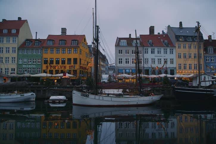 A view of a canal in a city with several colorful buildings with orange yellow and white walls and many sailboats parked in the water under an overcast sky