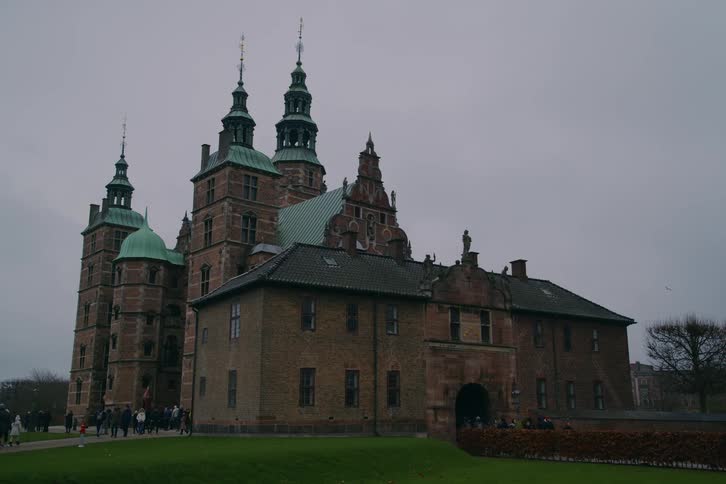 A historic european castle with green spires overlooks tourists walking on the lawn under gray sky