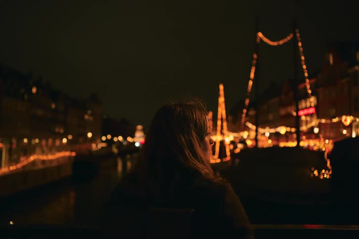 Woman sits near the river facing boats and streets decorated with lights in a residential area at night
