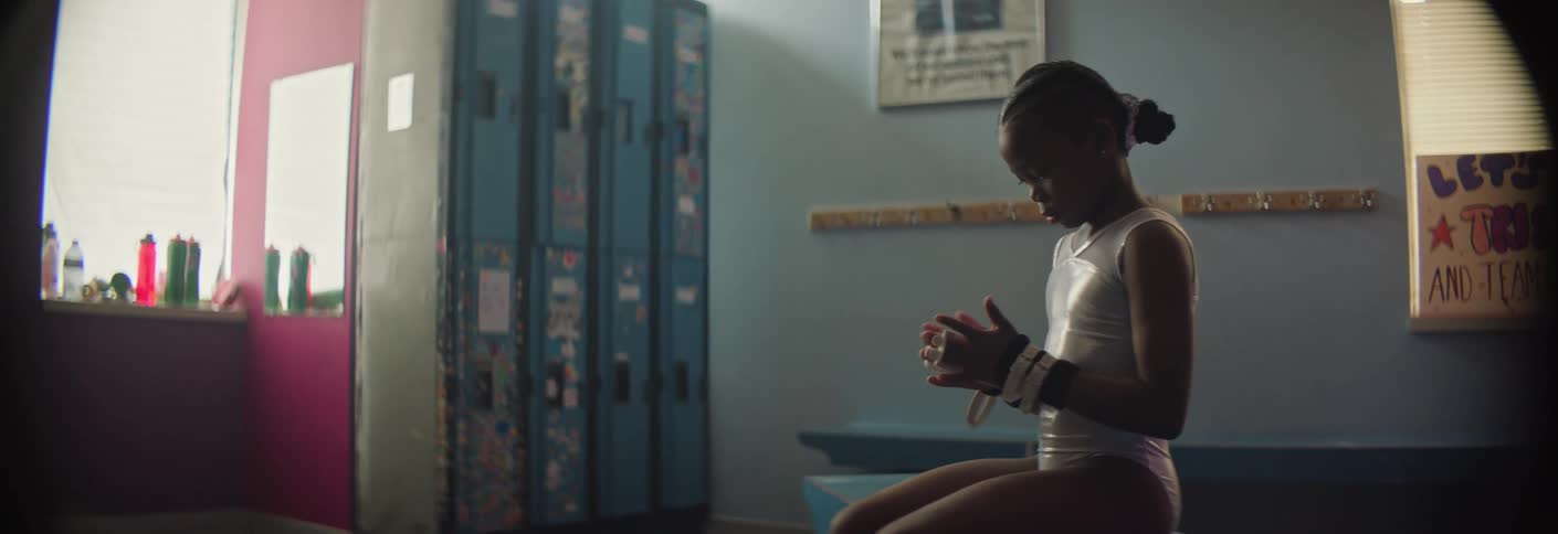 A young gymnast quietly prepares for practice while adjusting wrist guards in the locker room