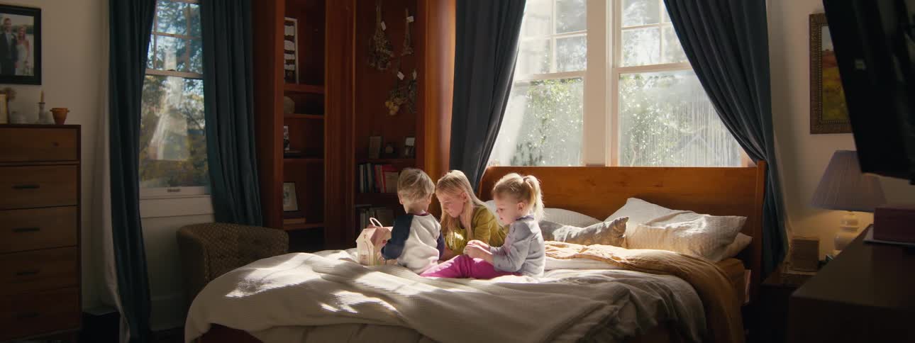 Three children sit on a bed interacting with toys in a bedroom lit by natural light