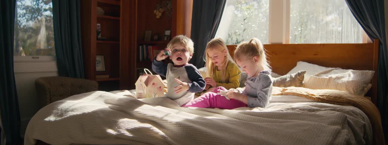 Three young siblings sit on a bed while playing with toys