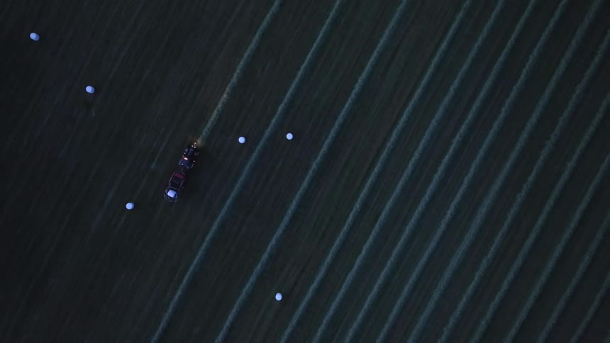 Topdown aerial view of a tractor working in a dark field at dusk driving past rows of harvested crops and scattered white round hay bales