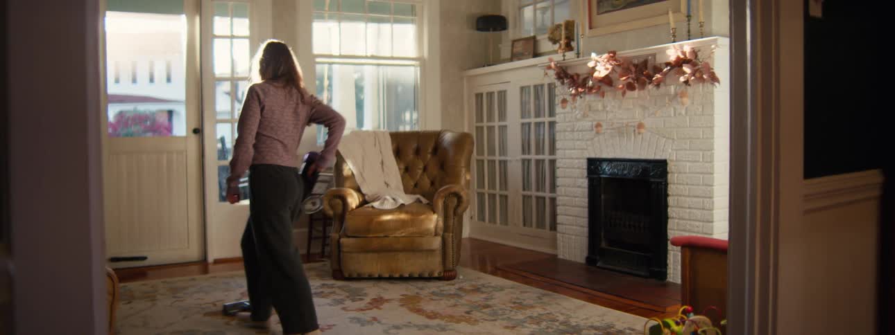 A woman vacuums an area rug in a living room while a toddler cries and reaches toward the camera in the foreground with his sister