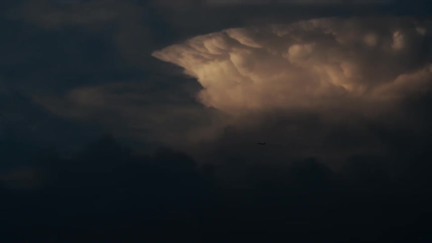 Airplane crosses a dark sky with a dense cloud illuminated in the background