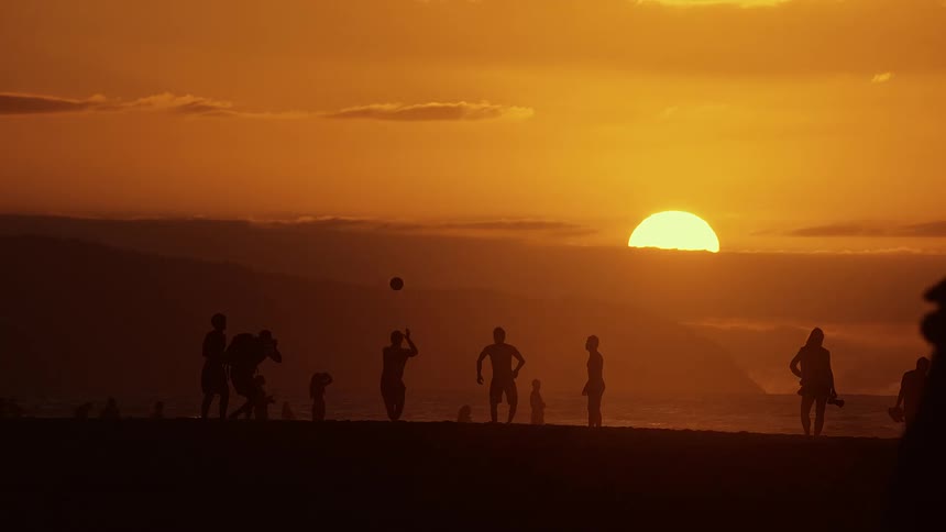 A group of people play with a soccer ball on a beach at sunset while others walk along the shore and the sun sits low on the horizon behind silhouettes of the crowd