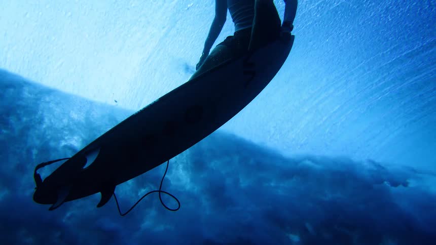 An underwater shot captures the silhouette of a surfer on a longboard as they paddle through churning white water and bubbles beneath the surface of a clear blue ocean wave