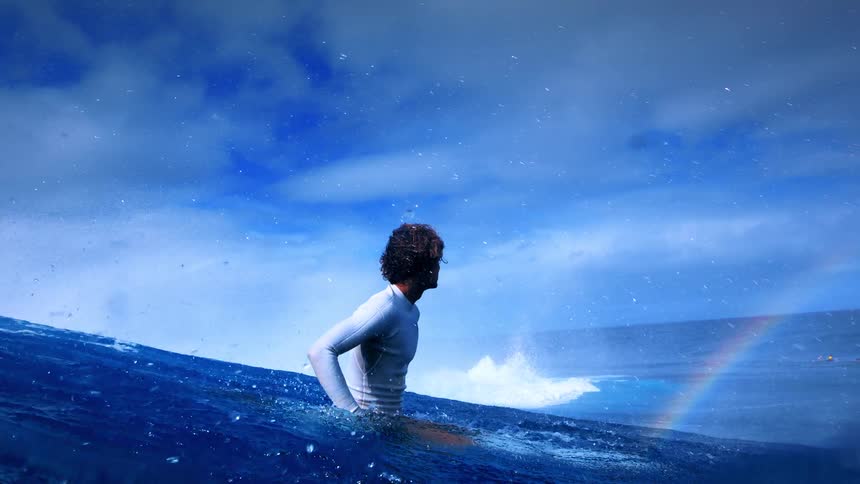 A man in a white longsleeve rashguard sits on his surfboard in deep blue water watching a wave break in the distance under a cloudy sky with a faint rainbow visible near the horizon