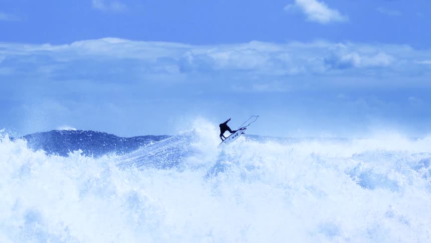 A surfer wearing a black wetsuit launches off a large wave performing a high aerial rotation before landing back into the churning white foam of the blue ocean under a cloudy sky