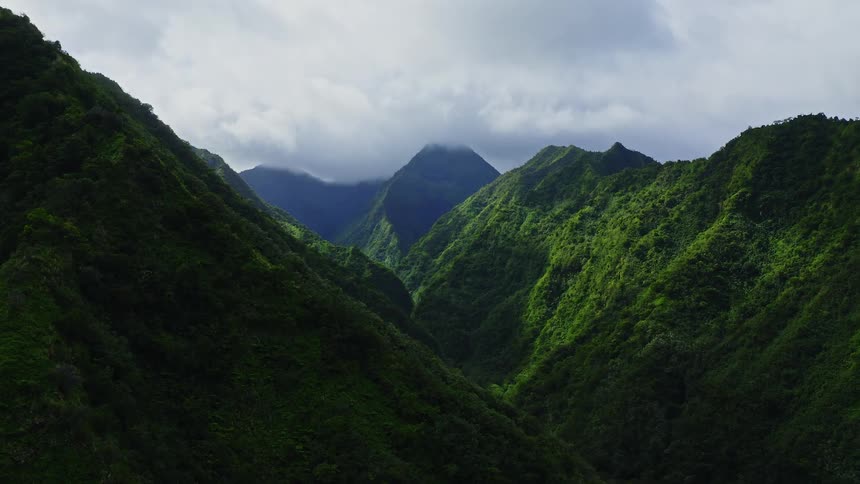 An aerial camera glides smoothly over a series of jagged emeraldgreen mountain ridges as thick mist and lowhanging clouds cling to the peaks and fill the deep verdant valleys under a heavy overcast sky