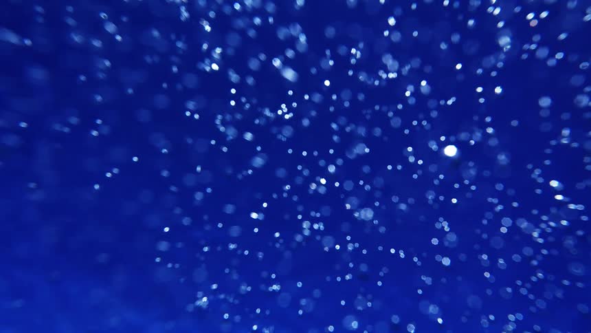 An abstract underwaterstyle shot shows countless shimmering white particles drifting through a deep blue field creating a calm ethereal atmosphere that feels like light suspended in water