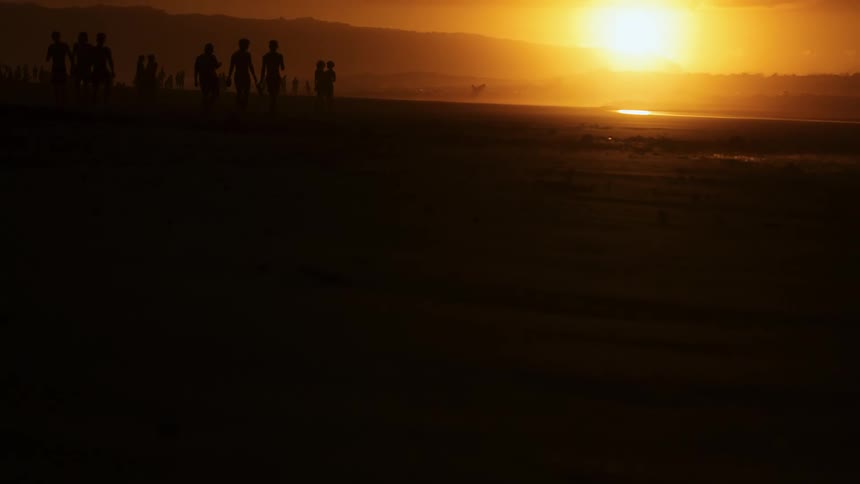 Silhouettes of people standing along the coast as the setting sun glows over a mountain in the distance