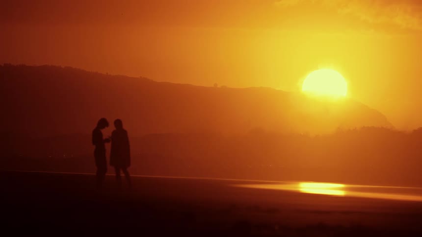 A man and woman stand in silhouette on a beach during a vibrant orange sunset looking down at their phones while waves gently wash onto the shore against a backdrop of dark hills and a large sun