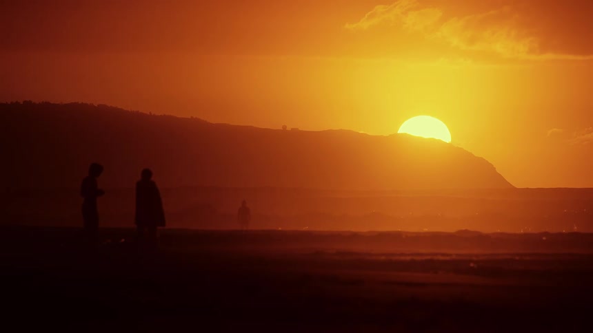 Silhouetted figures walk along a hazy beach as the large golden sun sets behind a distant mountain