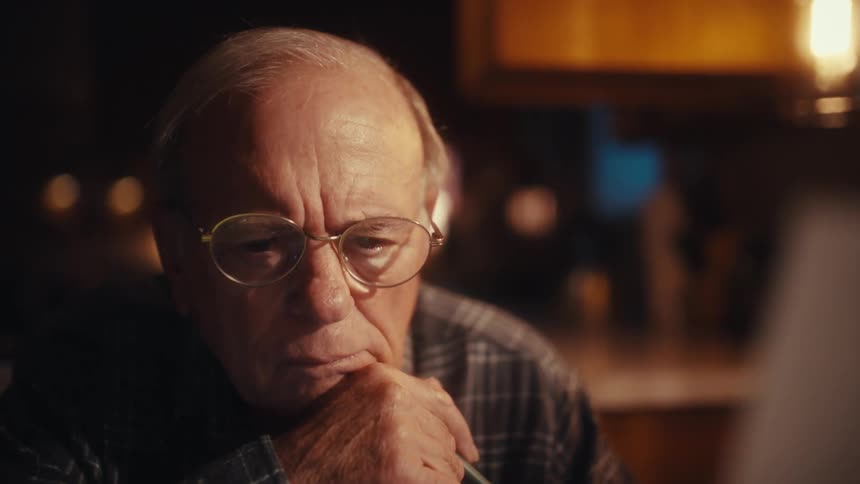 An elderly man in a plaid shirt is shown deeply focused while reviewing a document at his desk the video captures his concentrated expressions in closeup as he thoughtfully looks over the paperwork and his computer screen