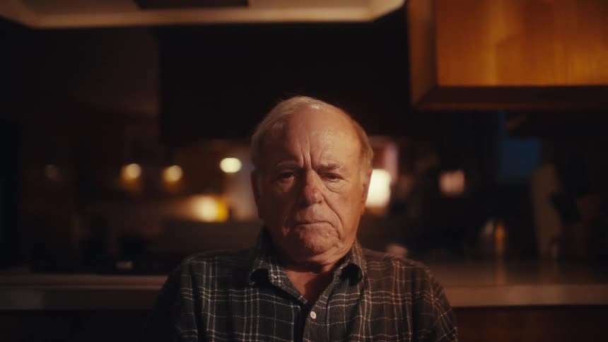 An elderly man sits in a dimly lit room with a somber expression his face showing deep contemplation as he looks away from a screen and rests his chin on his hands under warm ambient light