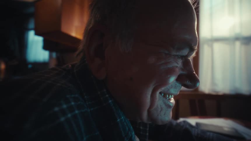 Man smiles as he sits at a table holding his glasses and looking at a laptop computer