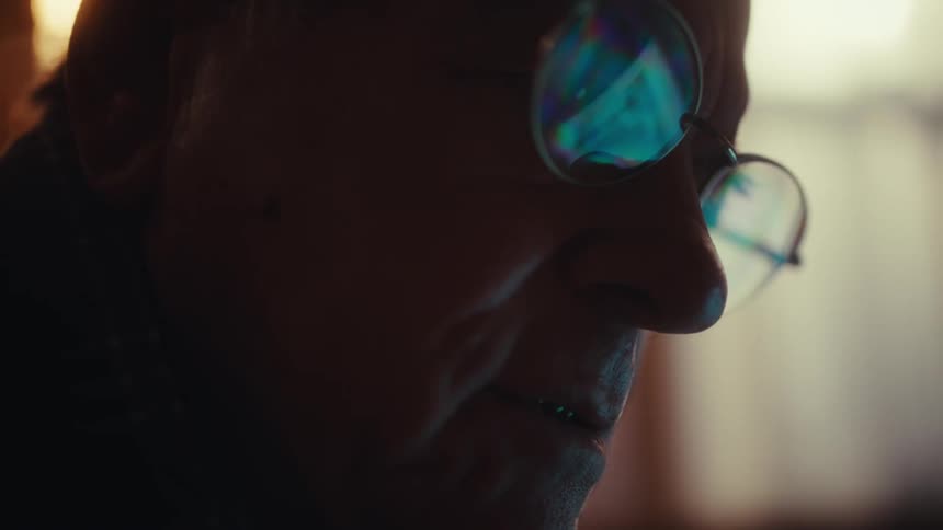 An elderly man wearing glasses looks down at a glowing screen in a dimly lit room his face reflecting the blue light before he breaks into a gentle smile while shifting his head slightly