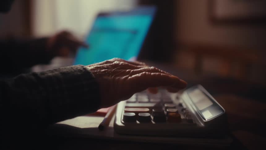 Elderly man references a computer while sitting at home and using a calculator