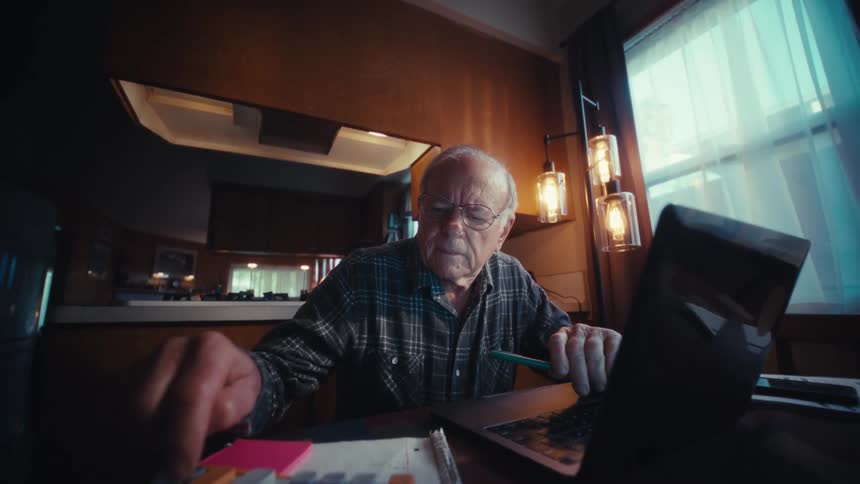 An elderly man sitting indoor operates a calculator while working on a laptop