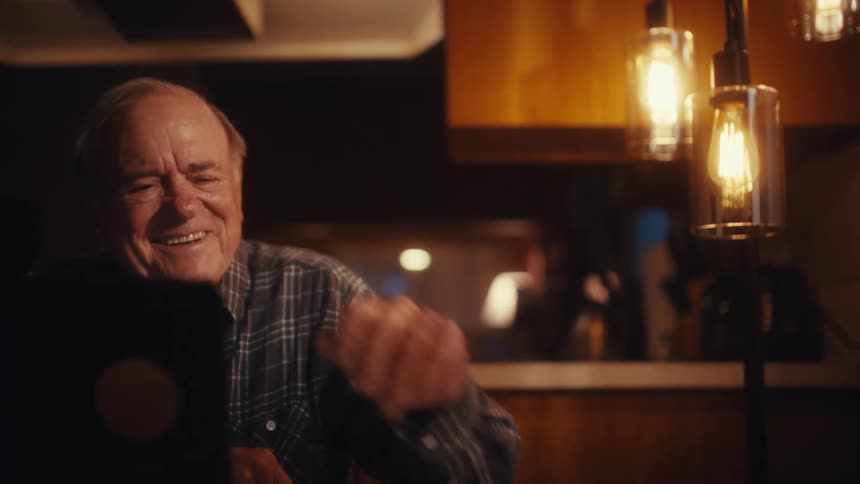 An elderly man in a plaid shirt sits at a desk carefully reading a white document before breaking into a wide heartfelt laugh and touching his face with genuine joy as warm decorative lightbulbs glow in the background of the dark room