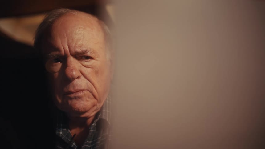 An elderly man in a plaid shirt sits at a desk under the warm glow of a lamp carefully reviewing a white document with a somber expression before leaning his head against his hand in quiet contemplation