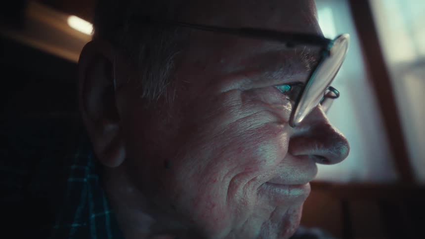An elderly man in a plaid shirt sits at a dining table using a laptop and a largebutton calculator then he puts on his glasses to read the screen and smiles briefly at the data