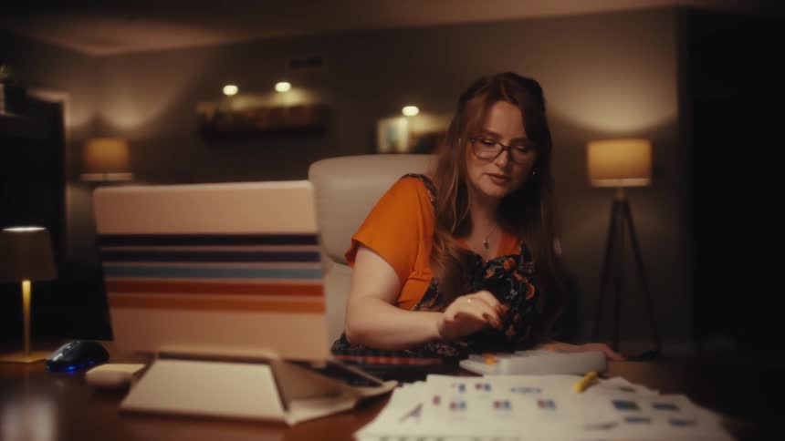 Young adult woman wearing glasses sitting and walking in front of a laptop computer while pushing buttons on a calculator over a table covered in paper sheets indoors during night time