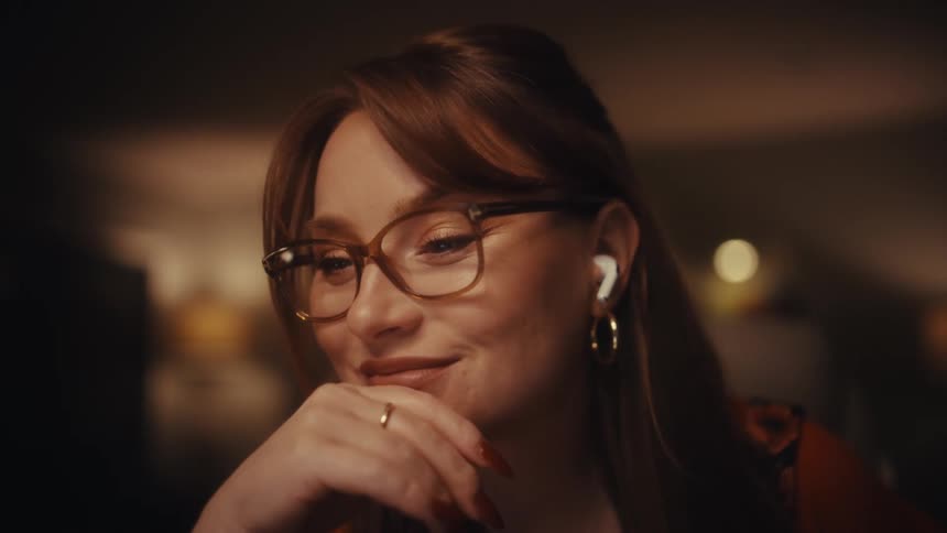 A young woman with red hair and glasses sits in a dimly lit room resting her chin on her hand while looking at a screen wearing wireless earbuds