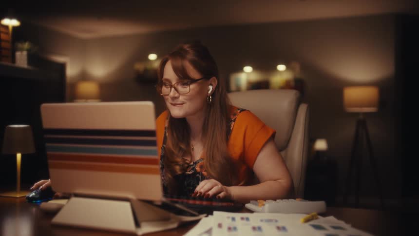 A woman sits at a desk working on a computer in a softly lit home office at night