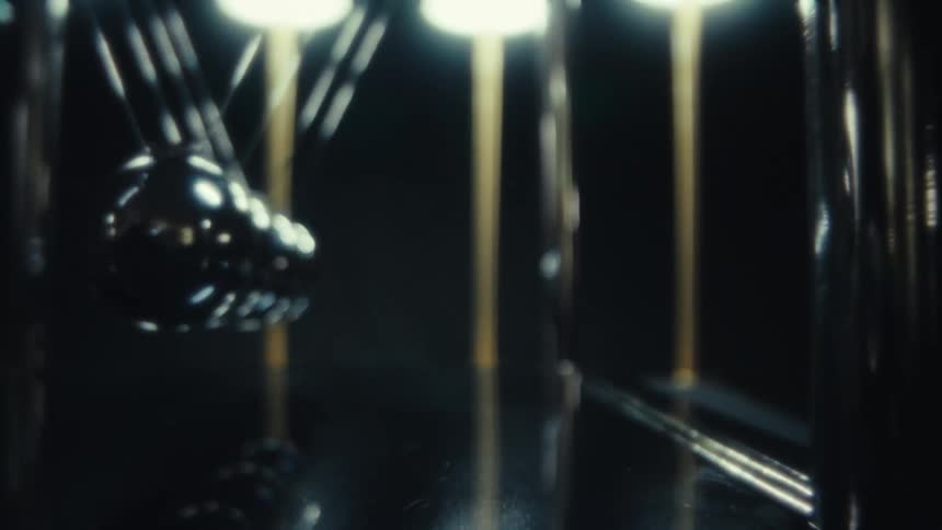 A metal sphere on a newtons cradle swings from the left and strikes a row of stationary chrome balls demonstrating the transfer of kinetic energy against a dark background with golden support pillars and soft overhead lighting
