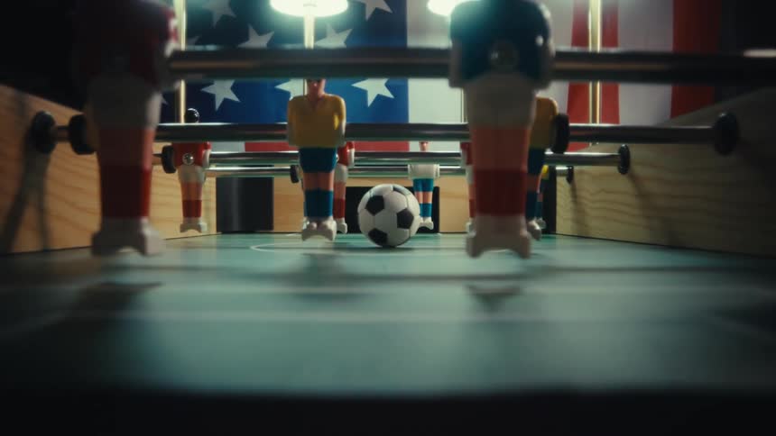 The video features a lowangle cinematic shot from inside a foosball table looking out toward the playersin the foreground a soccer ball sits stationary on the green pitch framed by the plastic legs of the foosball figures and an american flag han