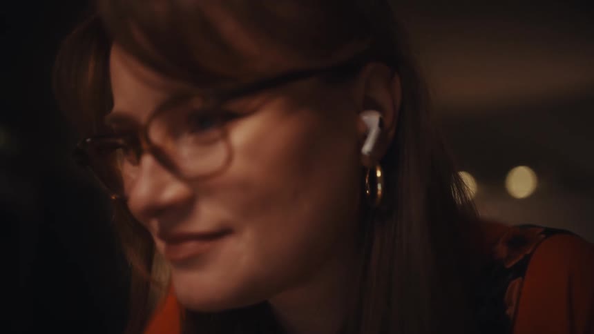 A closeup shot captures a woman with a manicured dark polish typing quickly on a mechanical keyboard with glowing orange keys before the camera pans up to reveal her wearing glasses and wireless earbuds as she works with a slight satisfied smile